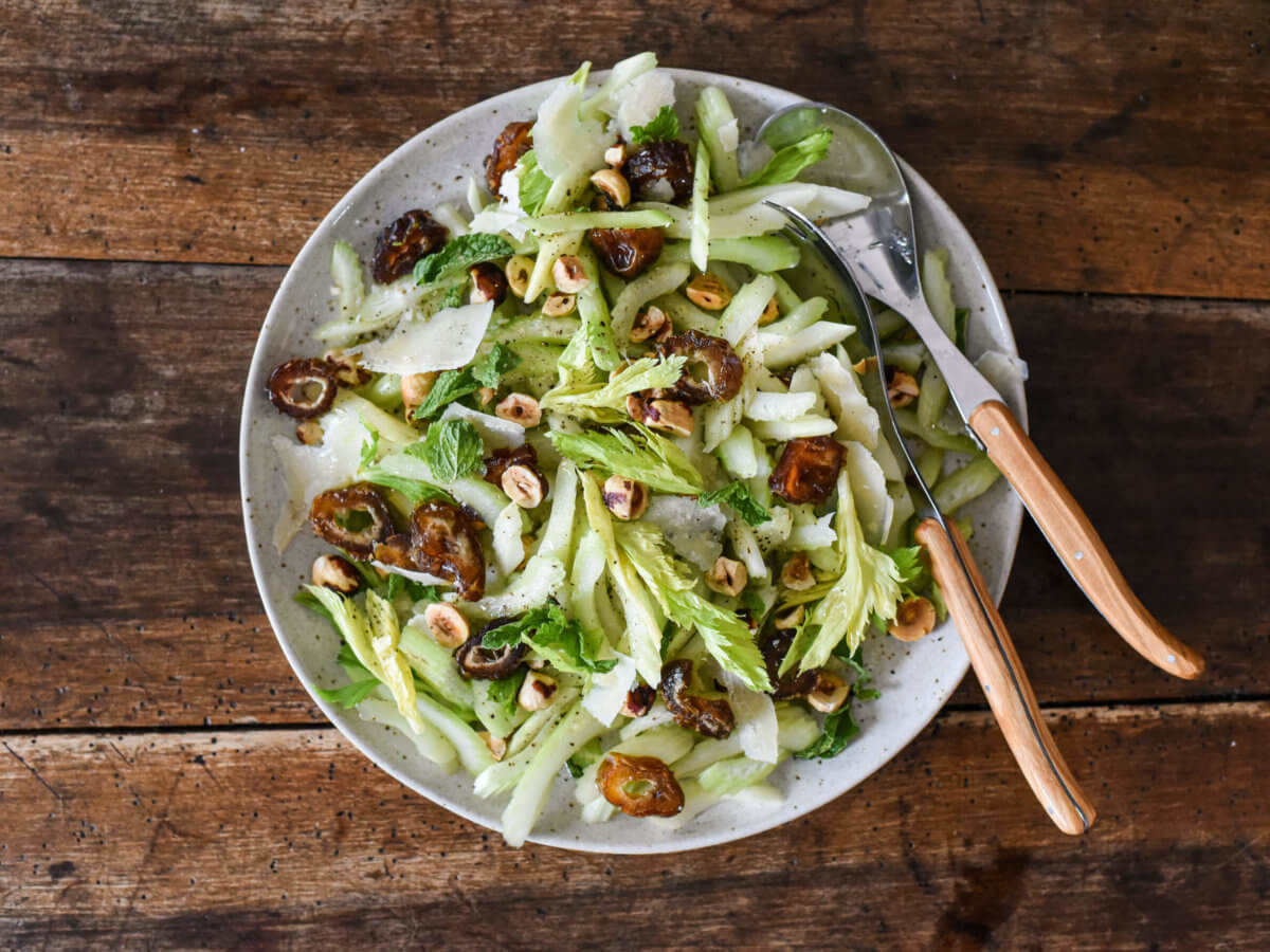 A plate of celery salad with dates, hazlenuts, parmesan cheese, and serving spoons